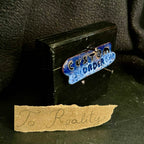 Finished cloisonné enamel 'Custom Order' piece on a black pine display stand with a handwritten 'To Reality' kraft paper label — completed custom enamel commission by A. Fraction, Vilnius
