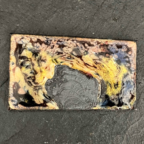 Fly Nest – original enamel miniature on copper,  up close view of the rectangular piece resting on dark slate. Swirling yellow, amber and black kiln-fired enamel around a burned grey central form. Handcrafted enamel art by A. Fraction.