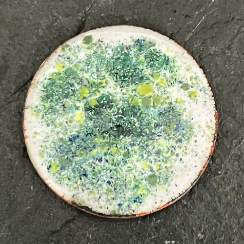 Existential Coin – original double-sided enamel art coin on copper, front view on dark slate. Round disc with mottled green, yellow and blue speckled enamel and a pale copper-edged rim. Handmade original artwork by A. Fraction.