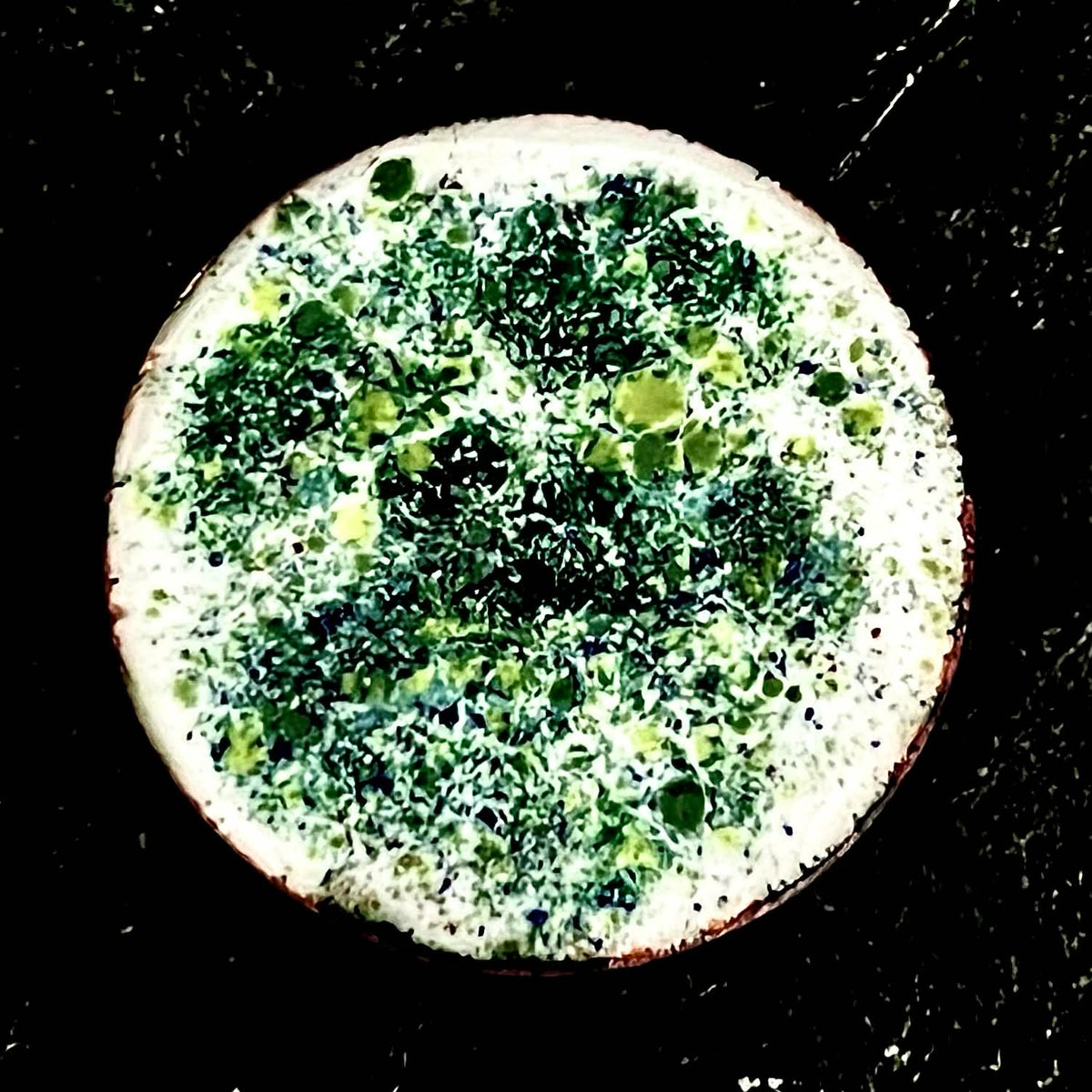 Existential Coin – original double-sided enamel art coin on copper, front view on dark slate. Round disc with mottled green, yellow and blue speckled enamel and a pale copper-edged rim. Handmade original artwork by A. Fraction.