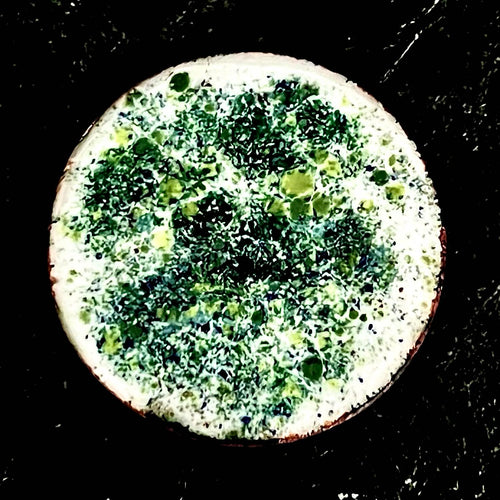 Existential Coin – original double-sided enamel art coin on copper, front view on dark slate. Round disc with mottled green, yellow and blue speckled enamel and a pale copper-edged rim. Handmade original artwork by A. Fraction.